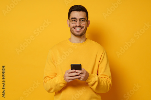 Guy wearing bright sweatshirt and eyeglasses, standing in front of camera with smartphone in hands, smiling happily, isolated on yellow background
