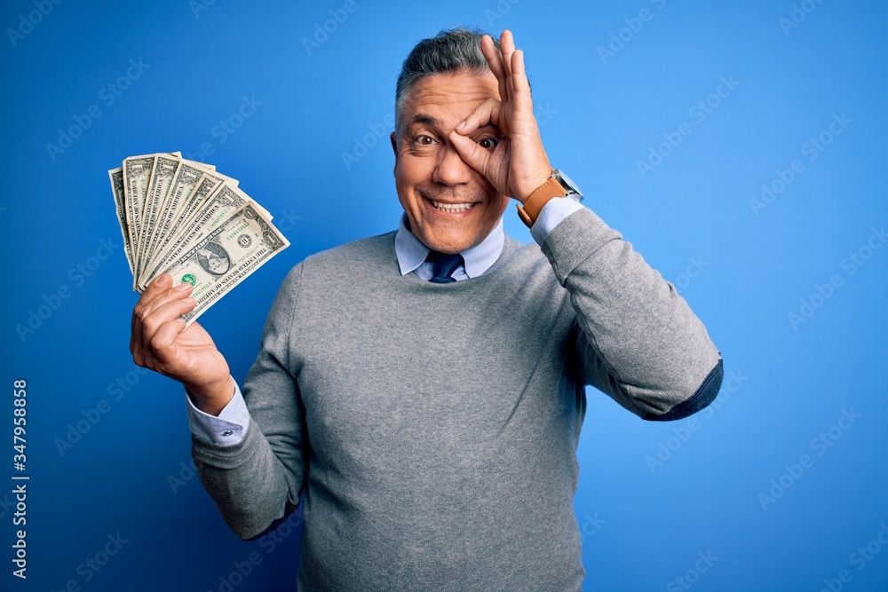 Middle age handsome grey-haired man holding bunch of dollars over blue background with happy face smiling doing ok sign with hand on eye looking through fingers Middle age handsome grey-haired man holding bunch of dollars over blue background with happy face smiling doing ok sign with hand on eye looking through fingers