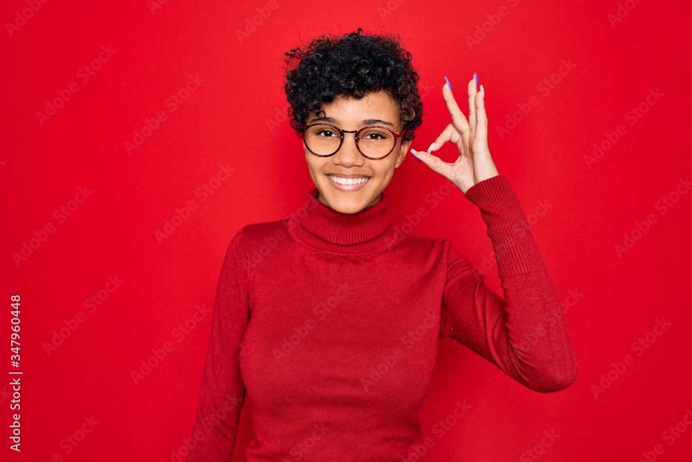Young beautiful african american afro woman wearing turtleneck sweater and glasses smiling positive doing ok sign with hand and fingers. Successful expression. Young beautiful african american afro woman wearing turtleneck sweater and glasses smiling positive doing ok sign with hand and fingers. Successful expression.