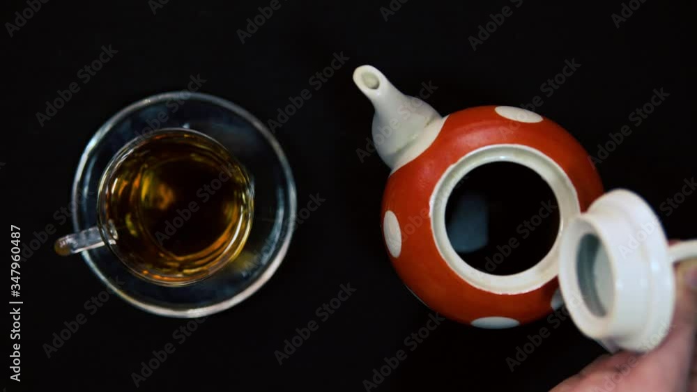 Tea ceremony. A male hand opens and closes the ceramic lid of a hot red polished white ceramic teapot next to a glass bowl for a bowl. Black background, oncoming light 4k Tea ceremony. A male hand opens and closes the ceramic lid of a hot red polished white ceramic teapot next to a glass bowl for a bowl. Black background, oncoming light 4k