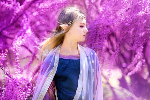 Beautiful romantic blonde girl with blue eyes posing in forest. Dreaming fairytale girl. Cosplay character. Portrait of elf girl with long ears touches pink flowers. Fairytale and fantasy work.