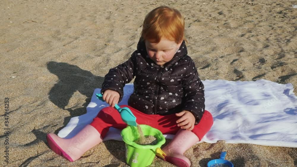 Beautiful red hair small kid in pink rubber boots is playing with plastic shovel and bucket on sand on the beach. Beautiful red hair small kid in pink rubber boots is playing with plastic shovel and bucket on sand on the beach.