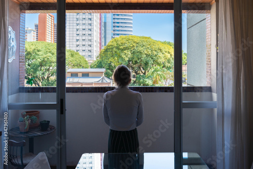 Mulher olhando o exterior, da porta de uma varanda, em dia ensolarado., durante a quarentena e isolamento social.