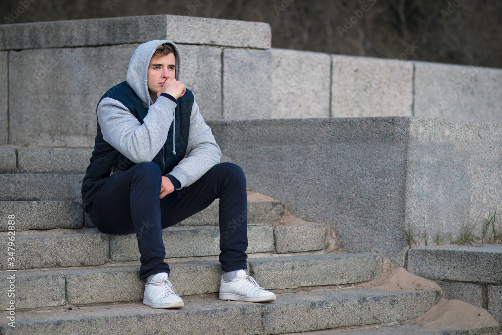 Sad upset depressed guy, young lonely frustrated man sitting on stairs in hood, suffering because of bad mood, thinking about future problems, looking into distance. Broken heart, failure concept. Sad upset depressed guy, young lonely frustrated man sitting on stairs in hood, suffering because of bad mood, thinking about future problems, looking into distance. Broken heart, failure concept.