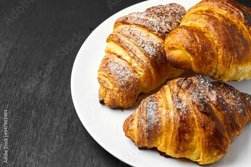 Top view on three fresh french buttery croissant on a white plate on a black background with copy space