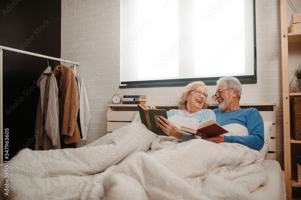 Happy elderly couple are lying in bed and reading favorite books while wearing glasses. Senior couple enjoying their time together. Happy elderly couple are lying in bed and reading favorite books while wearing glasses. Senior couple enjoying their time together.