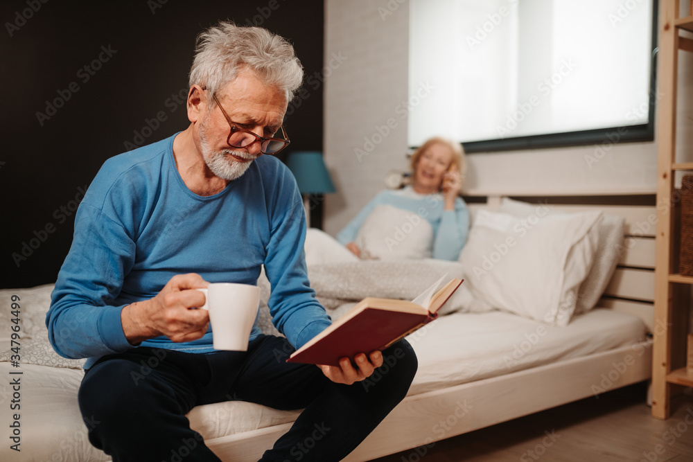Portrait of serious elderly man with grey hair and beard and with glasses reading book while drinking morning coffee. Blonde woman is talking over the phone while lying in bed behind him. Portrait of serious elderly man with grey hair and beard and with glasses reading book while drinking morning coffee. Blonde woman is talking over the phone while lying in bed behind him.