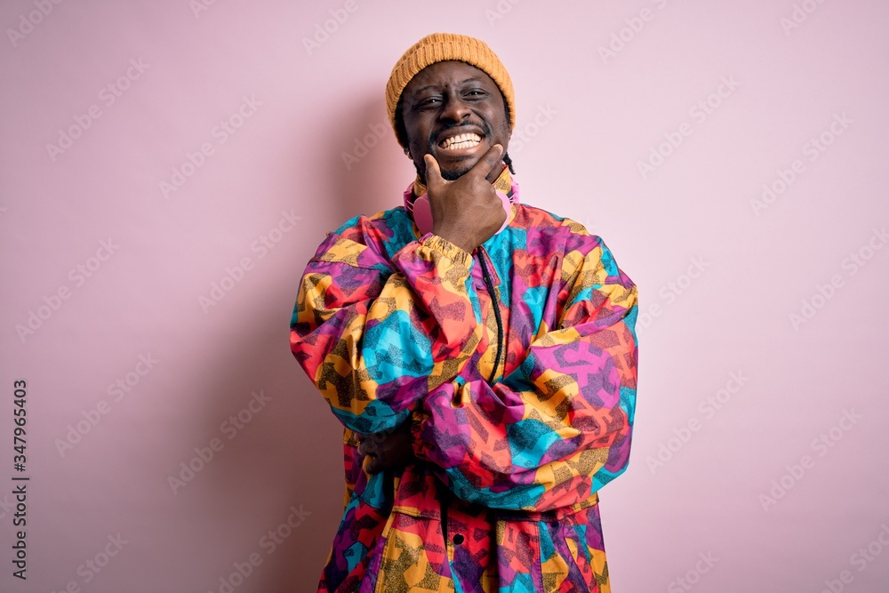 Young handsome african american man wearing colorful coat and cap over pink background looking confident at the camera smiling with crossed arms and hand raised on chin. Thinking positive.
