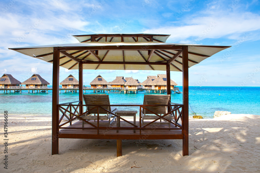 chairs on the sandy beach and houses over water of the blue sea. Polynesia.