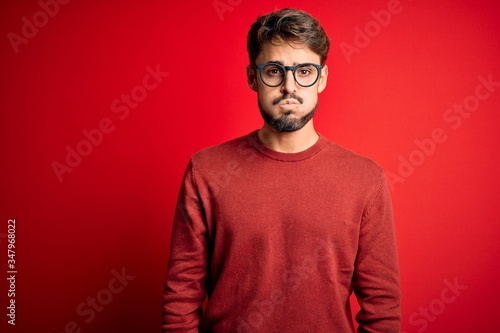 Young handsome man with beard wearing glasses and sweater standing over red background puffing cheeks with funny face. Mouth inflated with air, crazy expression.