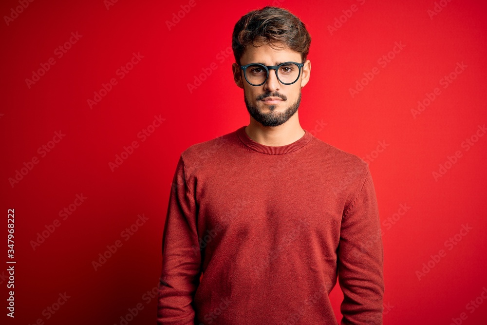 Young handsome man with beard wearing glasses and sweater standing over red background Relaxed with serious expression on face. Simple and natural looking at the camera. Young handsome man with beard wearing glasses and sweater standing over red background Relaxed with serious expression on face. Simple and natural looking at the camera.
