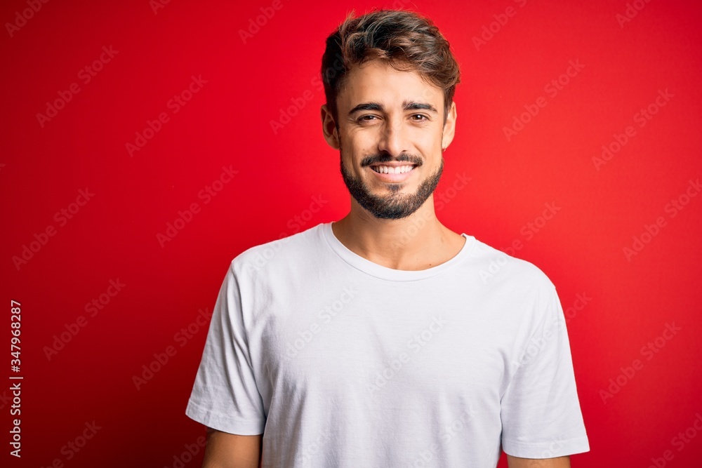 Young handsome man with beard wearing casual t-shirt standing over red background with a happy and cool smile on face. Lucky person. Young handsome man with beard wearing casual t-shirt standing over red background with a happy and cool smile on face. Lucky person.
