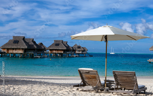 Two chairs on the sandy beach and houses over water of the blue sea. Polynesia.