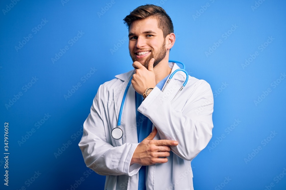 Young blond doctor man with beard and blue eyes wearing white coat and stethoscope looking confident at the camera with smile with crossed arms and hand raised on chin. Thinking positive. Young blond doctor man with beard and blue eyes wearing white coat and stethoscope looking confident at the camera with smile with crossed arms and hand raised on chin. Thinking positive.