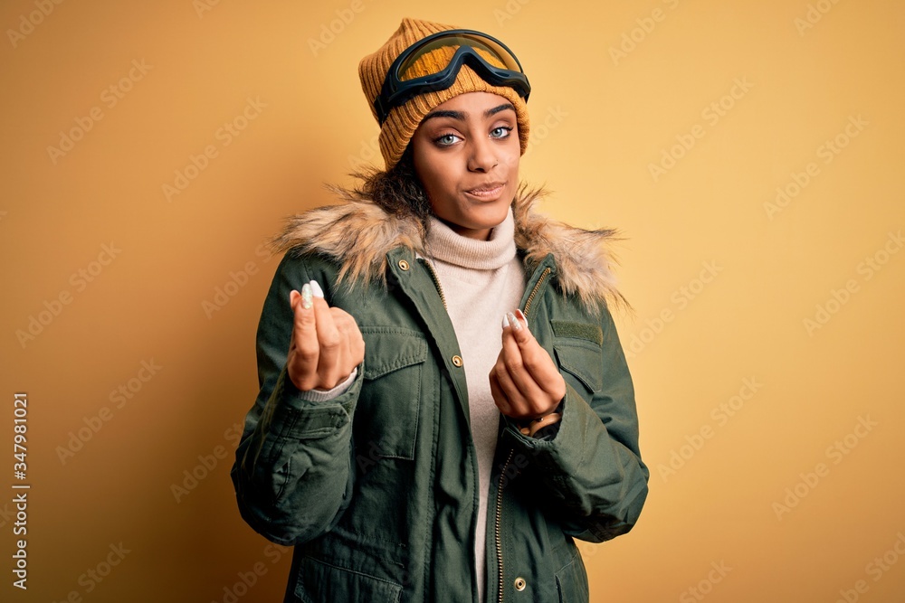 Young african american afro skier girl wearing snow sportswear and ski goggles doing money gesture with hands, asking for salary payment, millionaire business