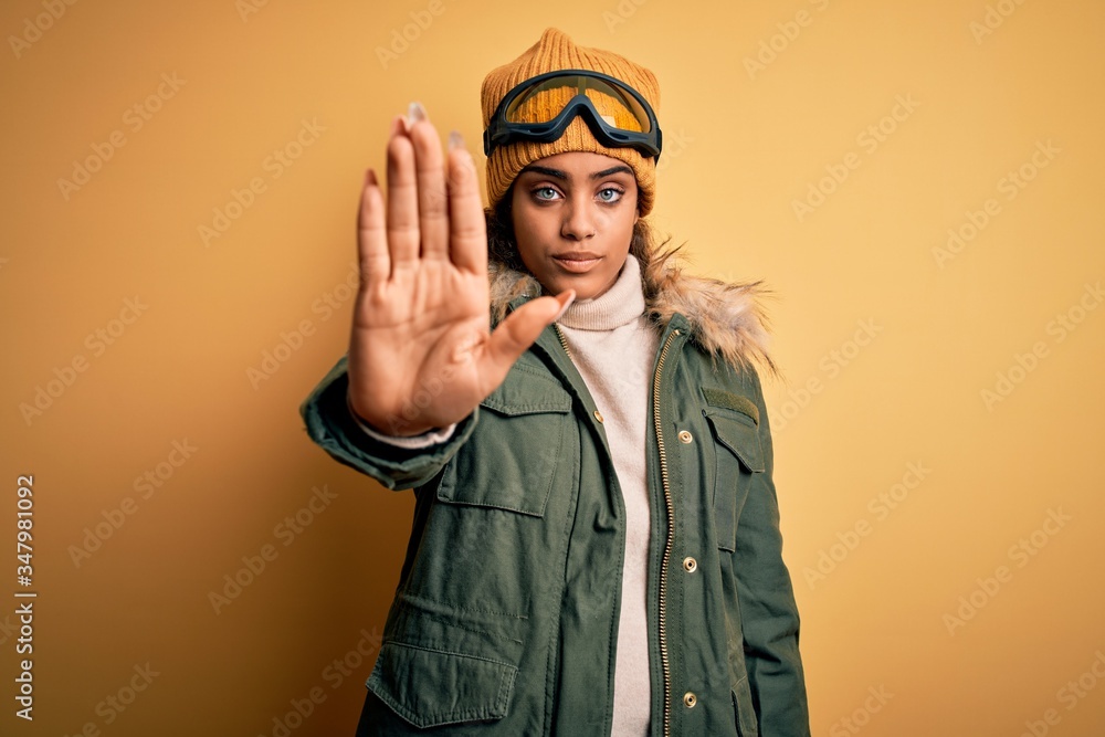 Young african american afro skier girl wearing snow sportswear and ski goggles doing stop sing with palm of the hand. Warning expression with negative and serious gesture on the face. Young african american afro skier girl wearing snow sportswear and ski goggles doing stop sing with palm of the hand. Warning expression with negative and serious gesture on the face.
