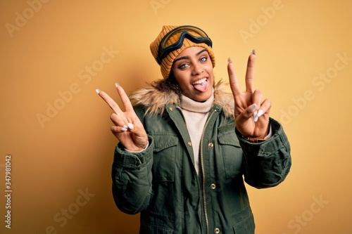 Young african american afro skier girl wearing snow sportswear and ski goggles smiling with tongue out showing fingers of both hands doing victory sign. Number two.
