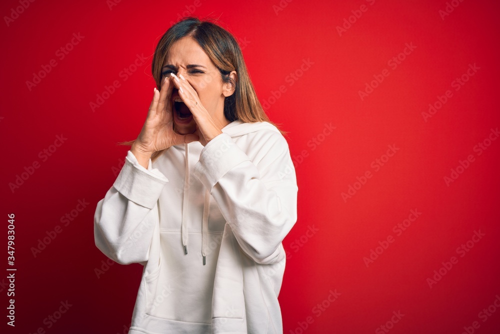 Middle age beautiful sportswoman wearing casual sweatshirt over isolated red background Shouting angry out loud with hands over mouth Middle age beautiful sportswoman wearing casual sweatshirt over isolated red background Shouting angry out loud with hands over mouth