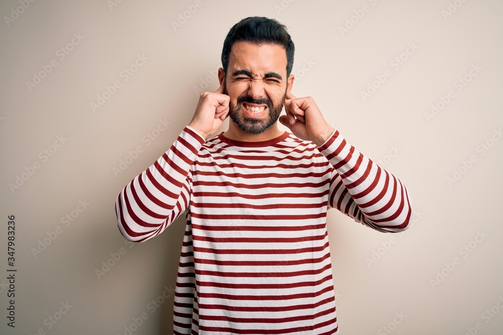 Young handsome man with beard wearing casual striped t-shirt standing over white background covering ears with fingers with annoyed expression for the noise of loud music. Deaf concept. Young handsome man with beard wearing casual striped t-shirt standing over white background covering ears with fingers with annoyed expression for the noise of loud music. Deaf concept.