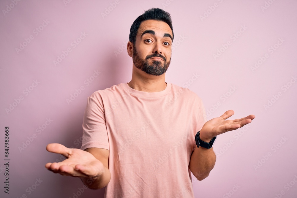 Young handsome man with beard wearing casual t-shirt standing over pink background clueless and confused expression with arms and hands raised. Doubt concept. Young handsome man with beard wearing casual t-shirt standing over pink background clueless and confused expression with arms and hands raised. Doubt concept.