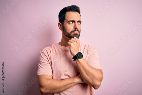 Young handsome man with beard wearing casual t-shirt standing over pink background with hand on chin thinking about question, pensive expression. Smiling with thoughtful face. Doubt concept.