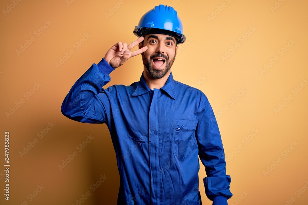 Mechanic man with beard wearing blue uniform and safety helmet over yellow background Doing peace symbol with fingers over face, smiling cheerful showing victory
