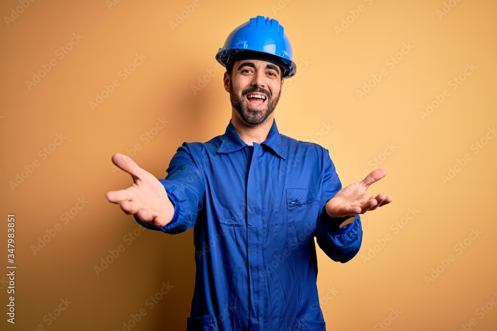 Mechanic man with beard wearing blue uniform and safety helmet over yellow background smiling cheerful offering hands giving assistance and acceptance.