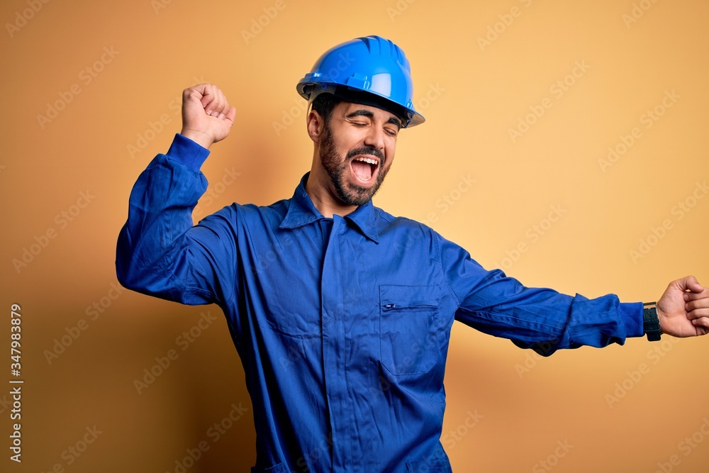 Mechanic man with beard wearing blue uniform and safety helmet over yellow background Dancing happy and cheerful, smiling moving casual and confident listening to music