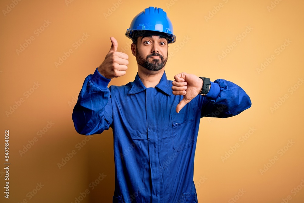 Mechanic man with beard wearing blue uniform and safety helmet over yellow background Doing thumbs up and down, disagreement and agreement expression. Crazy conflict