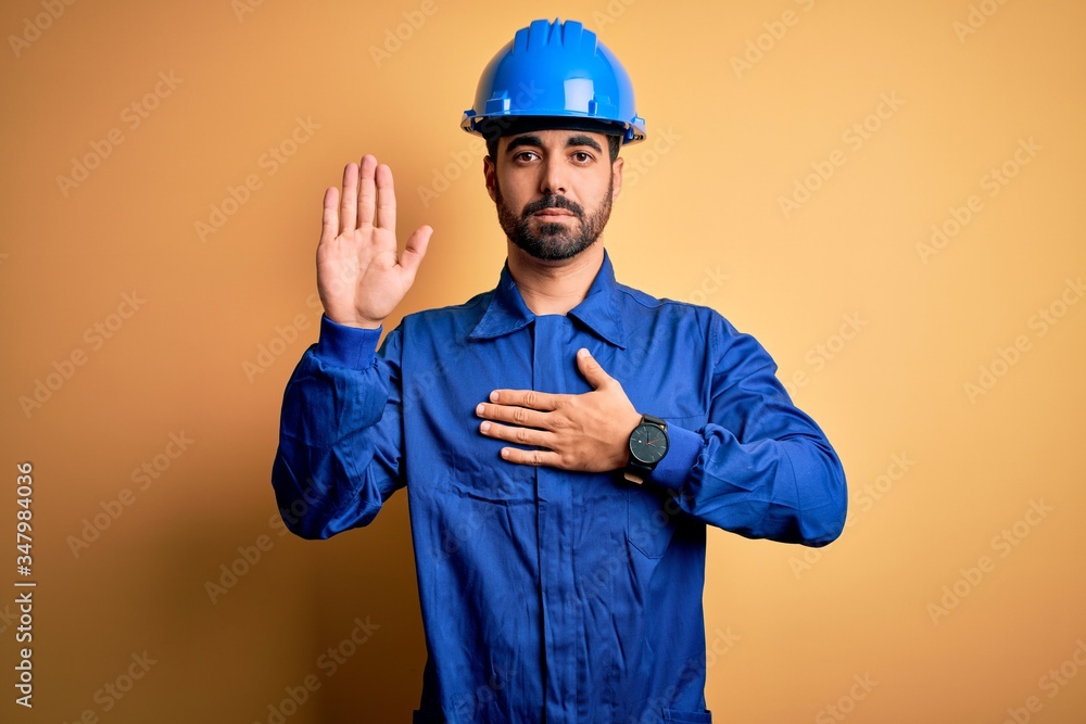 Mechanic man with beard wearing blue uniform and safety helmet over yellow background Swearing with hand on chest and open palm, making a loyalty promise oath