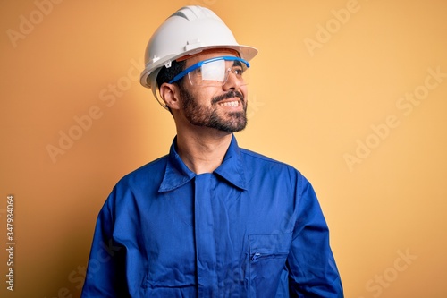 Mechanic man with beard wearing blue uniform and safety glasses over yellow background looking away to side with smile on face, natural expression. Laughing confident.