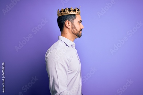 Young handsome man with beard wearing golden crown of king over purple background looking to side, relax profile pose with natural face with confident smile.
