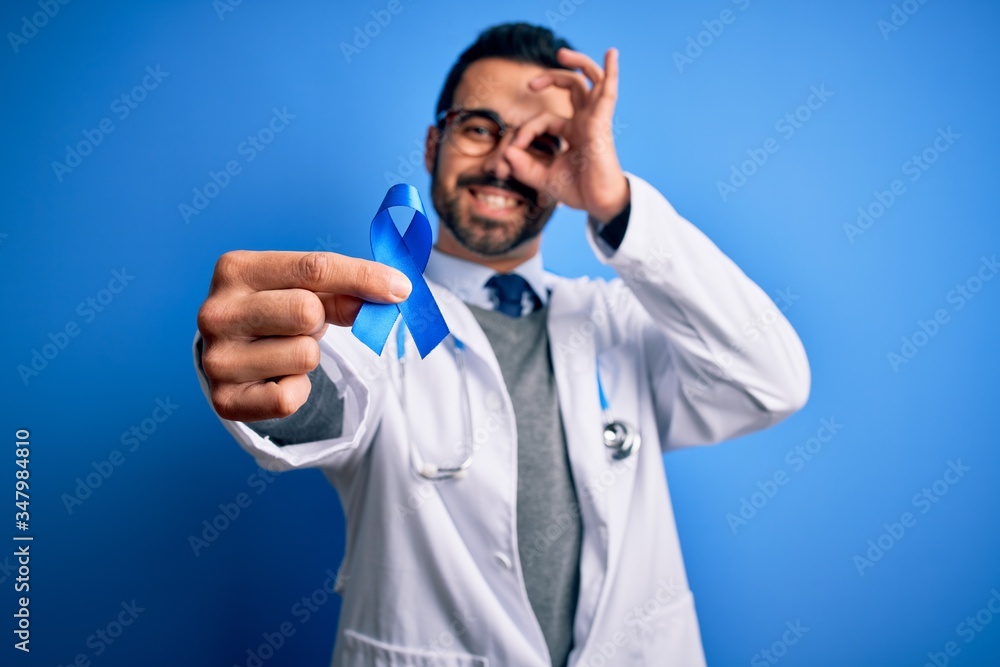 Young handsome doctor man with beard wearing stethoscope holding blue cancer ribbon with happy face smiling doing ok sign with hand on eye looking through fingers Young handsome doctor man with beard wearing stethoscope holding blue cancer ribbon with happy face smiling doing ok sign with hand on eye looking through fingers