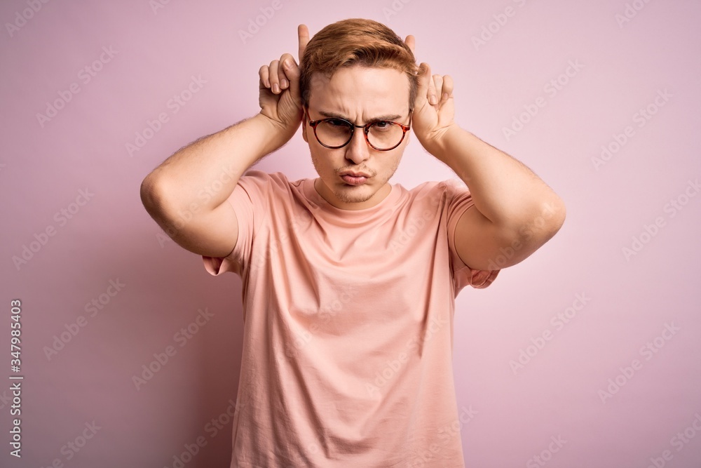 Young handsome redhead man wearing casual t-shirt standing over isolated pink background doing funny gesture with finger over head as bull horns Young handsome redhead man wearing casual t-shirt standing over isolated pink background doing funny gesture with finger over head as bull horns