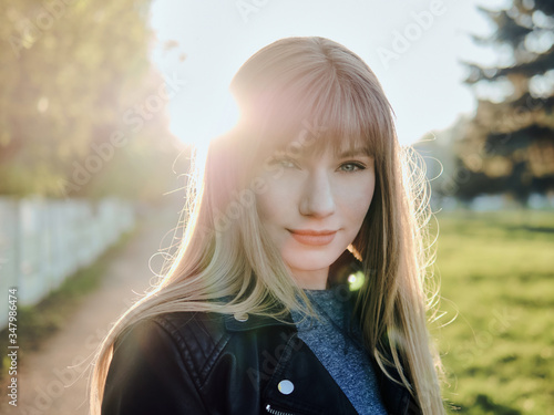 Close up headshot of adorable blonde haired charming smiling woman with blue eyes looking into camera and adorable smile in city park with bright sun flare evening
