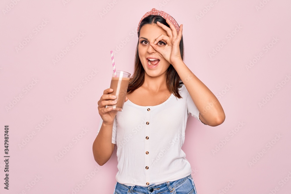 Young beautiful brunette woman drinking glass of chocolate beverage using straw smiling happy doing ok sign with hand on eye looking through fingers Young beautiful brunette woman drinking glass of chocolate beverage using straw smiling happy doing ok sign with hand on eye looking through fingers