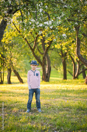 A boy in a hat and jeans stands in a blossoming apple orchard and holds his hands in his pockets. © Chendekova Liudmila