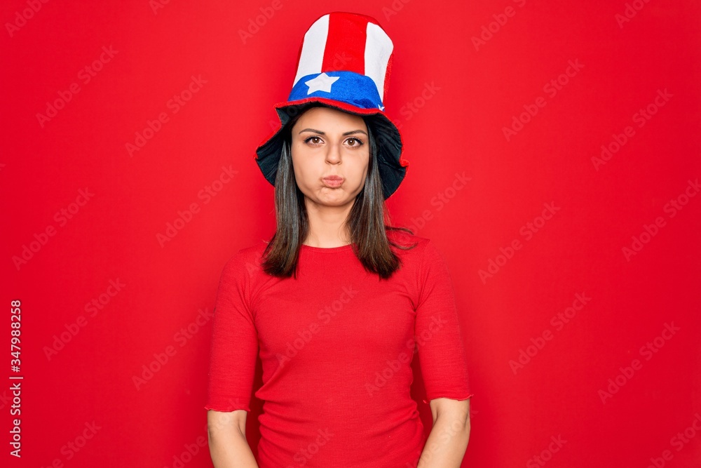 Young beautiful brunette woman wearing united states hat celebrating independence day puffing cheeks with funny face. Mouth inflated with air, crazy expression.