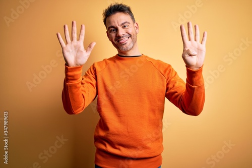 Young handsome man wearing orange casual sweater standing over isolated yellow background showing and pointing up with fingers number nine while smiling confident and happy.