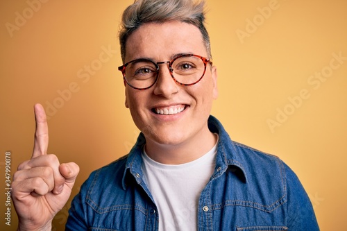 Close up of young handsome modern man wearing glasses and denim jacket over yellow background surprised with an idea or question pointing finger with happy face  number one