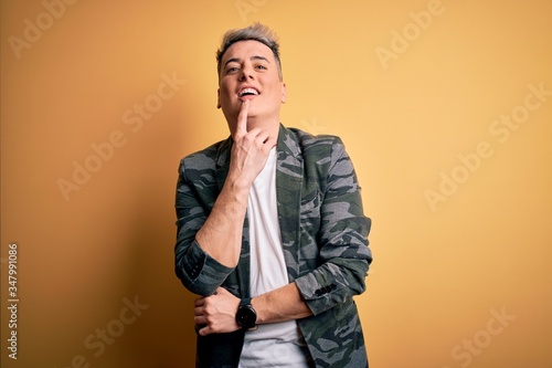 Young handsome modern man wearing business jacket standing over yellow isolated background looking confident at the camera with smile with crossed arms and hand raised on chin. Thinking positive.