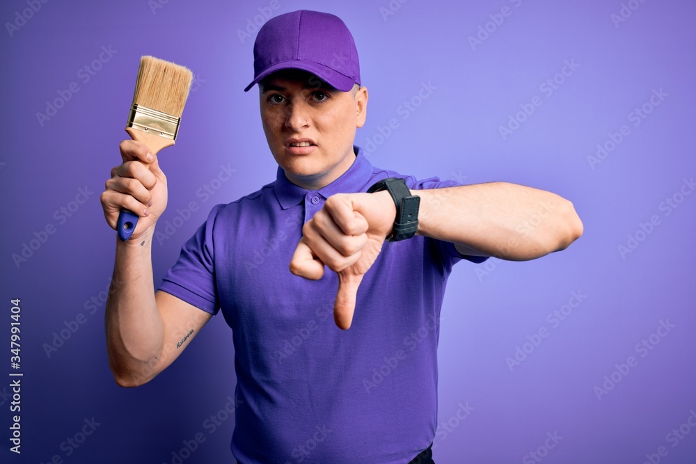 Young modern decorator painter man holding paint brush over purple background with angry face, negative sign showing dislike with thumbs down, rejection concept