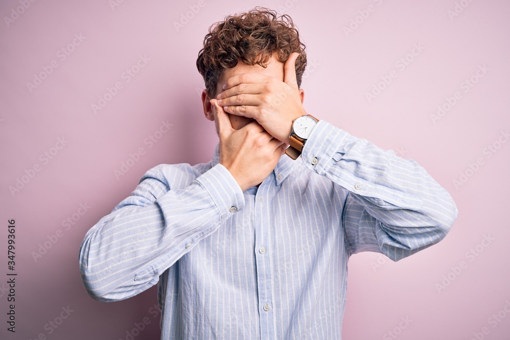 Young blond handsome man with curly hair wearing striped shirt over white background Covering eyes and mouth with hands, surprised and shocked. Hiding emotion Young blond handsome man with curly hair wearing striped shirt over white background Covering eyes and mouth with hands, surprised and shocked. Hiding emotion