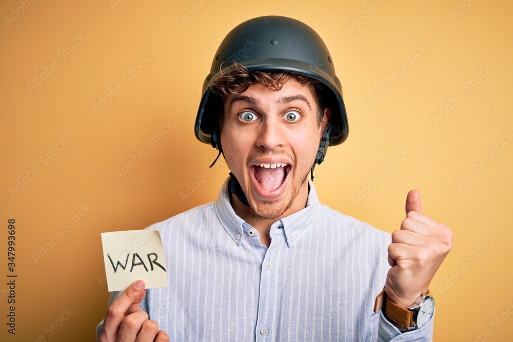 Young blond businessman with curly hair wearing helmet holding paper with war message screaming proud and celebrating victory and success very excited, cheering emotion