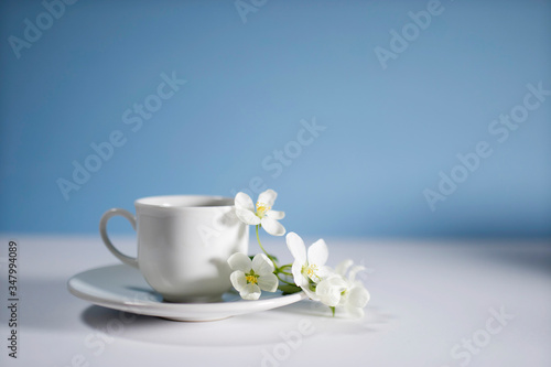 White cup of morning espresso with a blossoming apple tree branch on a stone surface of a table with reflections opposite blue background.