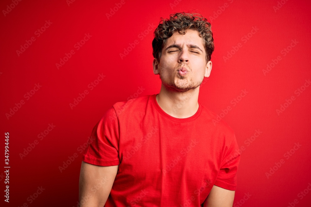 Young blond handsome man with curly hair wearing casual t-shirt over red background looking at the camera blowing a kiss on air being lovely and sexy. Love expression. Young blond handsome man with curly hair wearing casual t-shirt over red background looking at the camera blowing a kiss on air being lovely and sexy. Love expression.