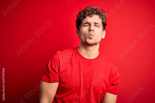Young blond handsome man with curly hair wearing casual t-shirt over red background looking at the camera blowing a kiss on air being lovely and sexy. Love expression.