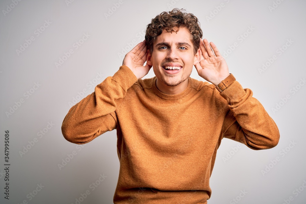 Young blond handsome man with curly hair wearing casual sweater over white background Trying to hear both hands on ear gesture, curious for gossip. Hearing problem, deaf Young blond handsome man with curly hair wearing casual sweater over white background Trying to hear both hands on ear gesture, curious for gossip. Hearing problem, deaf