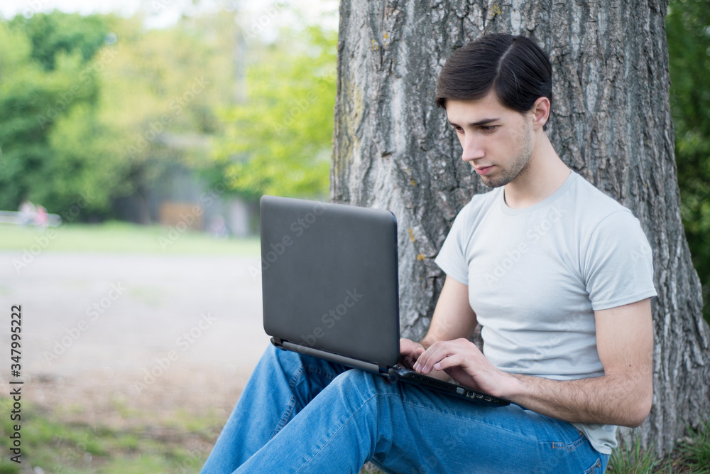 A young caucasian man sitting under a tree in a park and typing on a laptop. Remote work A young caucasian man sitting under a tree in a park and typing on a laptop. Remote work