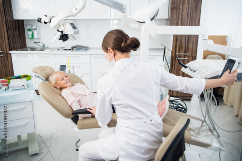 Skillful dentist is examining the teeth of a child. The woman is sitting near the girl trying to set up a contact with her to make her feel comfortable